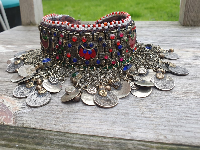 May include: A silver and gold choker necklace with red, blue, and green beads and coin-like charms. The necklace is made of metal and has a beaded band with a decorative design.