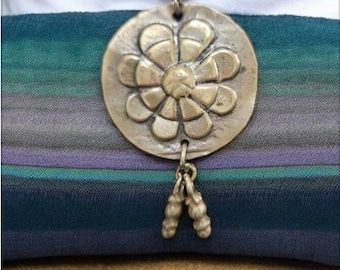 Könnte beinhalten: Eine Halskette aus Messing mit einem großen, runden Anhänger, der ein stilisiertes Blumendesign zeigt. Der Anhänger hat eine strukturierte Oberfläche und ist an einem braunen Band befestigt. Die Halskette hat eine kleine, dekorative Kette mit mehreren Perlen, die vom unteren Ende des Anhängers hängen.