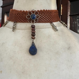 May include: A brown lace choker necklace featuring a central silver-toned flower charm with a blue stone inlay. A vertical drop hangs from the flower, consisting of four small brown wooden beads and a teardrop-shaped blue stone pendant.