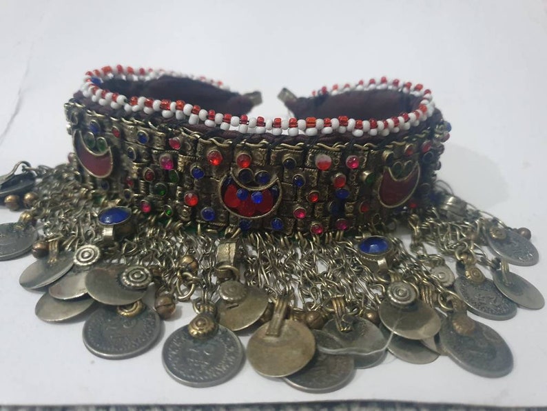 May include: A silver and gold necklace with red, blue, and green beads and a fringe of silver coins. The necklace is made of metal and has a beaded band with a decorative design.