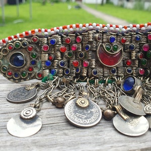 May include: A silver and gold beaded headband with red, blue, and green glass stones. The headband has a crescent moon design and is decorated with silver coins.