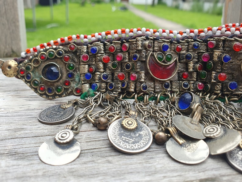 May include: A silver and gold beaded headband with red, blue, and green glass stones. The headband has a crescent moon design and is decorated with silver coins.