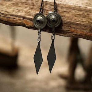 Op de afbeelding: Een paar zilverkleurige metalen oorbellen. Elke oorbel heeft een cirkelvormige bovenkant met een centraal gat en een ruitvormige hanger. De oorbellen hangen aan een dunne, donkere ketting en worden gepresenteerd op een stuk hout.