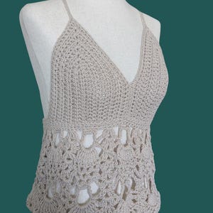 May include: A beige crochet halter top with a deep V-neck and delicate straps. The top features an openwork design at the waist, showcasing a scalloped edge. The handmade garment is displayed on a mannequin.