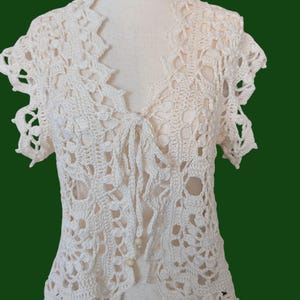 May include: An off-white crochet cardigan featuring an intricate floral pattern with short sleeves, a V-neckline, and a front tie closure. The garment is constructed with openwork stitching and includes two small round beads at the bottom of the tie.