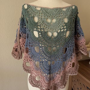 May include: A handmade crochet shawl in a gradient of colors, including blue, green, and pink. The openwork design features circular patterns and a scalloped edge. The shawl is displayed on a mannequin.
