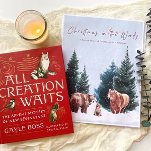 May include: Two hardback books and a lit candle in a glass jar are displayed on a cream-coloured cloth. One book is titled "All Creation Waits" with illustrations of a fox, bird, and squirrel. The other book is titled "Christmas in the Woods" with illustrations of bears and trees.