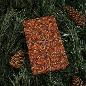 May include: A wrapped gift with a brown, embossed floral pattern, tied with twine, rests on a bed of green pine needles. Two pine cones are visible. The gift wrap has a detailed, textured design.