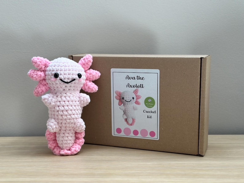 May include: A pink crocheted axolotl amigurumi plushie with black eyes. The plushie is packaged in a brown box with a label that reads "Ava the Axolotl Crochet Kit".