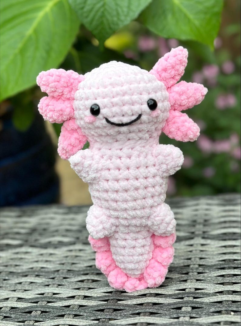 May include: A pink crocheted axolotl plush toy with black eyes and a smiling mouth. The toy is standing on a woven surface.