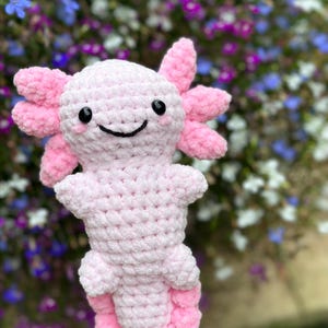 May include: A pink crocheted axolotl plush toy with a smiling face. The toy is made of soft yarn and has a fluffy texture.