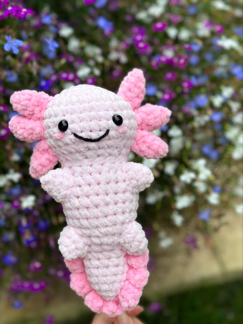 May include: A pink crocheted axolotl plush toy with a smiling face. The toy is made of soft yarn and has a fluffy texture.