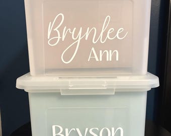 May include: Two stacked rectangular plastic storage bins with lids. The top bin is translucent white with the white text Brynlee Ann. The bottom bin is a light blue color with the white text Bryson Charles printed on the front surface.
