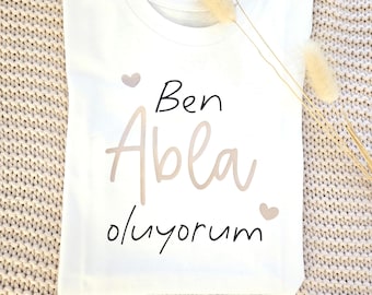 May include: A white t-shirt featuring the text "Ben Abla oluyorum" in black and beige script, with heart graphics. The shirt is folded on a textured beige surface, with dried flowers. The text is in Turkish.