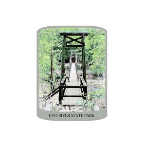 May include: A wooden suspension bridge spans a river, surrounded by lush green trees. The bridge has a dark wooden frame and a wooden walkway. The text "ENO RIVER STATE PARK" is at the bottom of the image.