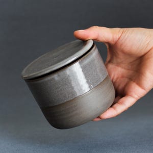 Op de afbeelding: Een cilindrische keramische houder met deksel, voorzien van een tweekleurig ontwerp. Het bovenste gedeelte en het deksel hebben een glanzende, lichtgrijze afwerking, terwijl het onderste gedeelte een matte, donkergrijze afwerking met textuur heeft.