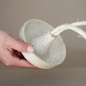 Może przedstawiać: Nakrapiany ceramiczny klosz lampy wiszącej z kremowym sznurem. Oprawa jest trzymana przez dłoń, ukazując jej zaokrąglony kształt i nakrapianą powierzchnię. Sznur jest gruby i skręcony.