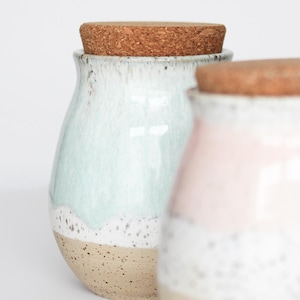 Può includere: Due vasi in ceramica con coperchi in sughero. Il vaso a sinistra ha una glassa azzurra sulla metà superiore, una fascia bianca maculata e una base beige maculata. Il vaso a destra ha una glassa rosa, una fascia bianca maculata e una base beige maculata.