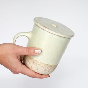 Può includere: Una tazza in ceramica verde chiaro con coperchio. La metà inferiore della tazza è beige macchiato. Ha un manico e un coperchio abbinato con un piccolo pomello. La tazza è tenuta in diagonale.