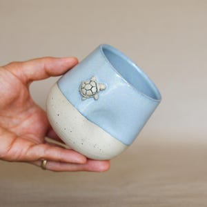 Könnte beinhalten: Ein Keramikbecher mit zweifarbigem Design, bestehend aus einem glänzend hellblauen oberen Teil und einer gesprenkelten cremeweißen Basis. Eine kleine, plastisch geformte Schildkrötenfigur ist an der Außenseite des blauen Bereichs angebracht.
