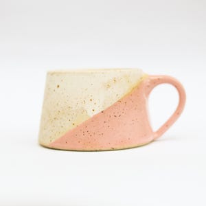 Può includere: Una tazza in ceramica fatta a mano con un design unico. La tazza è divisa diagonalmente, con una sezione color crema macchiettata e una sezione rosa. Ha un manico arrotondato e un corpo leggermente angolato.