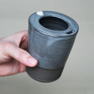 Könnte beinhalten: Ein handgefertigter, dunkelgrauer Keramik-Reisebecher mit einer einzigartigen ovalen Öffnung und einem kleinen, weißen Akzent. Der Becher hat ein zweifarbiges Design mit dunklerem Boden und hellgrauem Oberteil. Der Becher wird von einer Person gehalten.