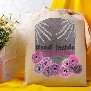May include: Beige tote bag with a gray tombstone design featuring skeleton hands with pink nails, the words "Dead inside," and pink and green floral eye graphics. The text "BRITTKRUGERDESIGNS" is also visible.