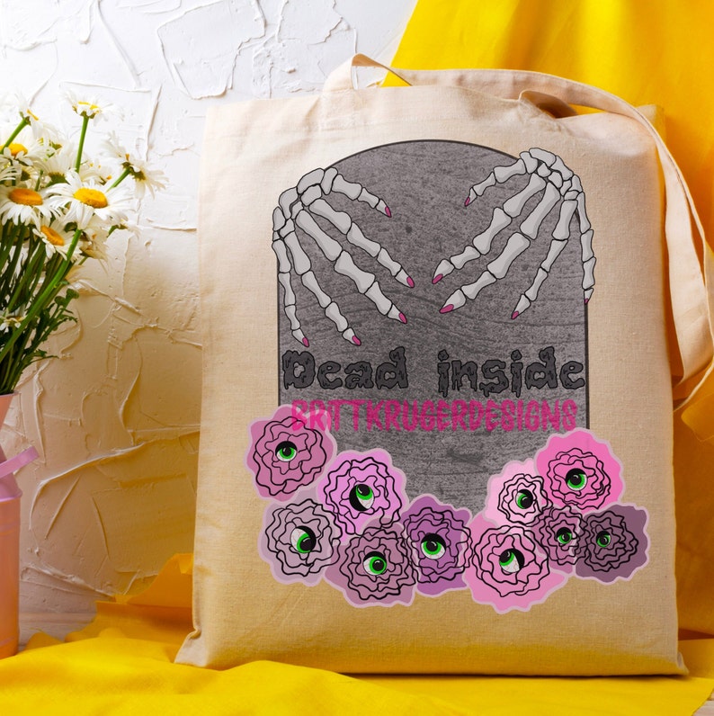 K&ouml;nnte beinhalten: Beige Tragetasche mit einem grauen Grabstein-Design mit Skeletth&auml;nden mit rosa N&auml;geln, den Worten "Dead inside" und rosa und gr&uuml;nen floralen Augengrafiken. Der Text "BRITTKRUGERDESIGNS" ist ebenfalls sichtbar.