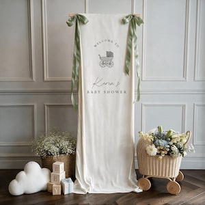 May include: A vertical cream-coloured fabric banner hanging from a wooden rod with sage green ribbon bows. The banner features a grey baby carriage graphic and the text "WELCOME TO Karin's BABY SHOWER" printed in dark grey lettering.