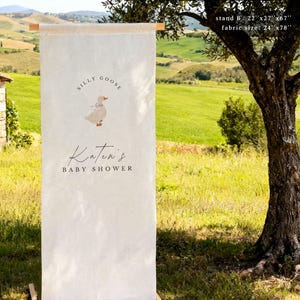 May include: A vertical off-white linen sign suspended from a wooden rod, featuring a graphic of a goose with a blue bow, the text "SILLY GOOSE", "Katen's", and "BABY SHOWER". The sign dimensions are listed as stand 22"x27"x67" and fabric 24"x78".