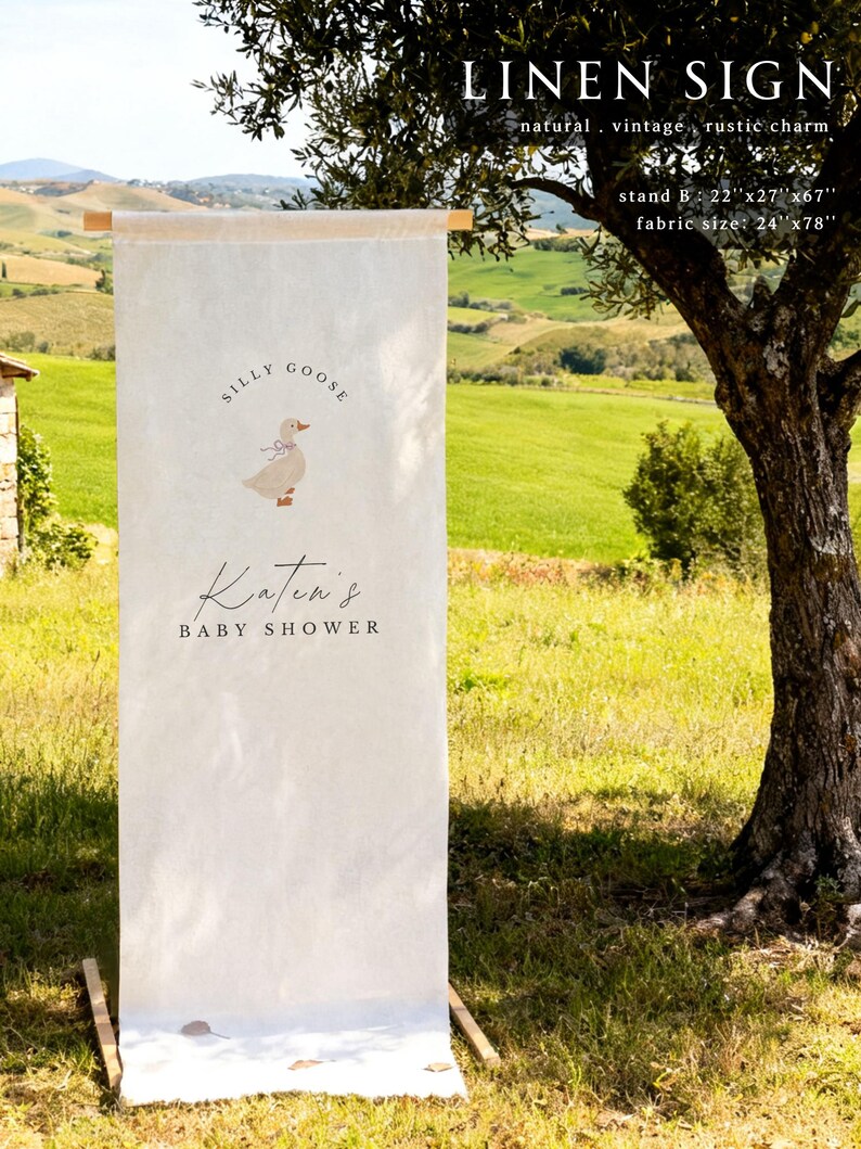 May include: A vertical off-white linen sign suspended from a wooden rod, featuring a graphic of a goose with a blue bow, the text "SILLY GOOSE", "Katen's", and "BABY SHOWER". The sign dimensions are listed as stand 22"x27"x67" and fabric 24"x78".