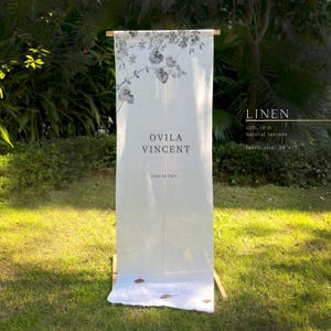 Może przedstawiać: Biały lniany baner z kwiatowym wzorem oraz imionami "OVILA VINCENT" i datą "June 18, 2024". Baner ma wymiary 61 cm x 191 cm i jest prezentowany na drewnianym stojaku w ogrodzie.