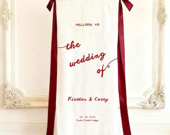 Może przedstawiać: Pionowy baner ślubny z białej tkaniny z ciemnoczerwonym napisem "WELCOME TO the wedding of Kirsten & Corey 19.06.2025 Lake Creek Lodge". Baner jest ozdobiony dwiema długimi, ciemnoczerwonymi satynowymi wstążkami zawiązanymi w kokardy na górze.
