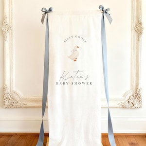 May include: A vertical off-white linen sign featuring the text SILLY GOOSE above an illustration of a small goose wearing a blue bow. Below the goose, the text Katen's BABY SHOWER is printed. The fabric is suspended by two long blue ribbons.