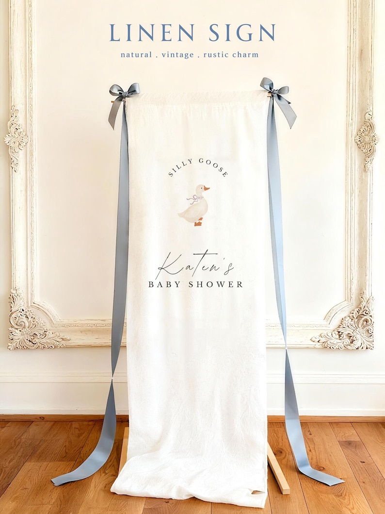 May include: A vertical off-white linen sign featuring the text SILLY GOOSE above an illustration of a small goose wearing a blue bow. Below the goose, the text Katen's BABY SHOWER is printed. The fabric is suspended by two long blue ribbons.