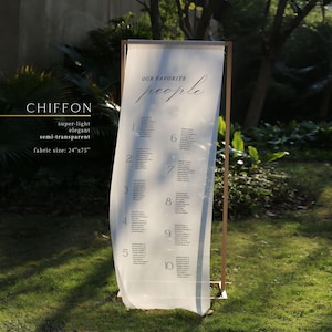 May include: A semi-transparent chiffon fabric wedding seating chart with the text "OUR FAVORITE people". The fabric is 24" x 75" and is displayed on a gold metal frame. Numbers 1-10 are visible on the chart.