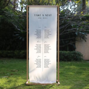 Könnte beinhalten: Ein Hochzeits-Sitzplan in einem Holzrahmen. Der Text "TAKE A SEAT" steht oben, mit den Namen der Gäste in Spalten darunter. Der Hintergrund ist eine verschwommene Außenszene.