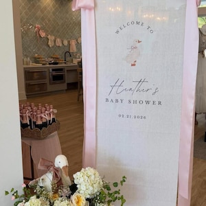 May include: A tall, vertical white fabric banner featuring a printed illustration of a duck with a pink bow, the text "WELCOME TO", "Heather's", "BABY SHOWER", and the date "02.21.2026". The banner is accented with long, light pink satin ribbons.
