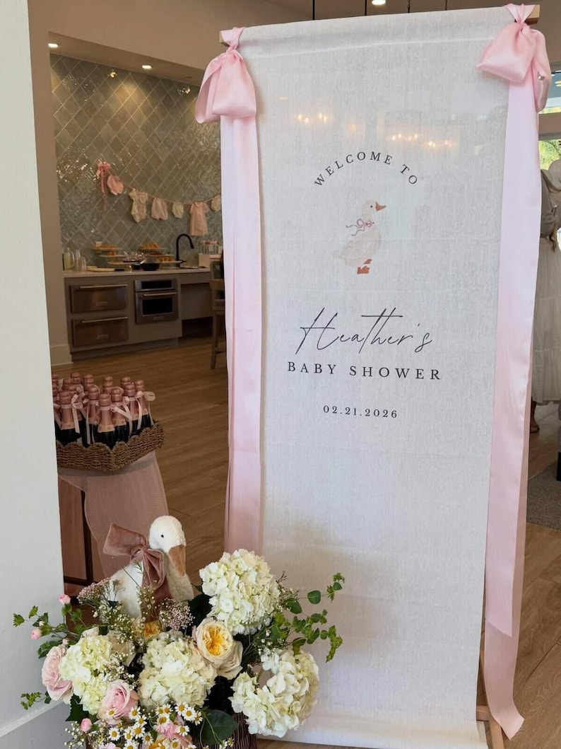May include: A tall, vertical white fabric banner featuring a printed illustration of a duck with a pink bow, the text "WELCOME TO", "Heather's", "BABY SHOWER", and the date "02.21.2026". The banner is accented with long, light pink satin ribbons.