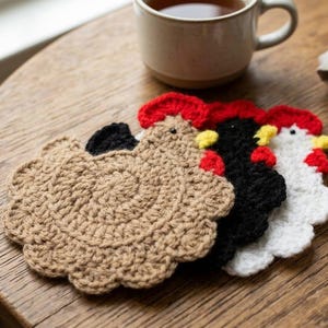 May include: Three crocheted coasters shaped like chickens, each featuring a distinct color scheme. One is tan, one is black, and one is white. Each coaster has a red comb, a small yellow beak, and a dark eye, with a scalloped edge design.