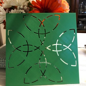 Peut inclure: Panneau décoratif carré vert avec un motif floral découpé. Le panneau présente un motif symétrique de lignes courbes et de formes en croix. En arrière-plan, un bouquet de fleurs colorées.