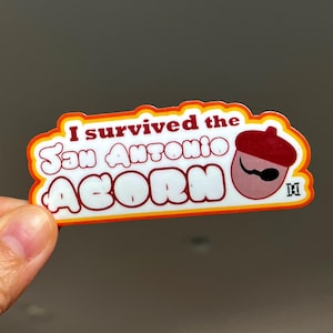 May include: A white sticker with orange and red borders. The sticker says "I survived the San Antonio Acorn" in a retro font. A cartoon acorn wearing a red beret and a mustache is on the right side of the sticker.