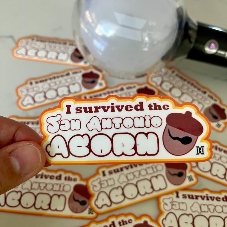 May include: A sticker with a white background and orange, red, and brown accents. The sticker says "I survived the San Antonio ACORN" with a cartoon acorn wearing a brown hat and a mustache.