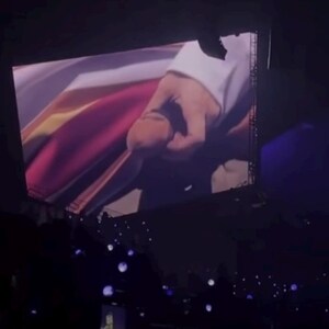 May include: A large screen displaying a close-up of a hand holding a colorful fabric. The screen is in a dark room with a crowd of people in the foreground.