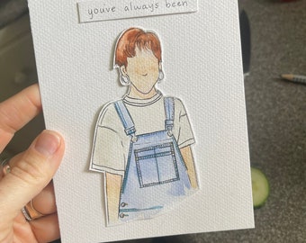 May include: Hand-drawn greeting card with a watercolor illustration of a person wearing blue overalls and a white t-shirt. The card has the text "you've always been" above the illustration. The card is held by a hand.