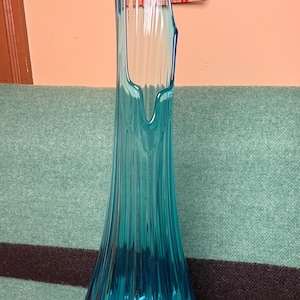 May include: A tall, turquoise-colored glass vase with a flared base and a narrow, fluted body. The vase has a unique, asymmetrical top and is approximately 12 inches tall. The ribbed texture catches the light, creating visual interest. A vintage decorative piece.
