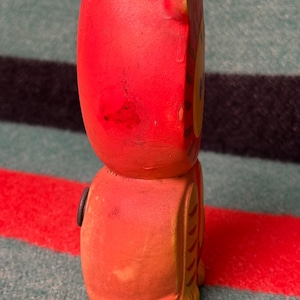 May include: A red and orange ceramic tiger figurine with a hollow top opening. The tiger features a yellow face, painted brown stripes on the side, and a small circular black detail on the side of its body. The surface shows signs of wear.