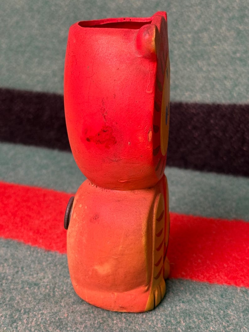 May include: A red and orange ceramic tiger figurine with a hollow top opening. The tiger features a yellow face, painted brown stripes on the side, and a small circular black detail on the side of its body. The surface shows signs of wear.