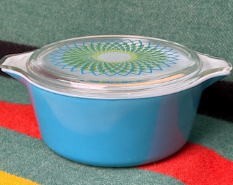 May include: A vintage turquoise Pyrex casserole dish with a clear glass lid. The lid features a green and blue floral design. The dish has two handles and is set against a colorful striped background.