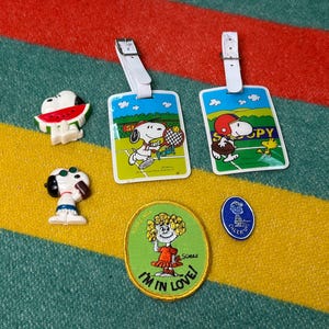 May include: A collection of Snoopy-themed items, including two luggage tags with Snoopy playing tennis and baseball, two Snoopy figurines, a patch with the text "I'M IN LOVE!", and a blue pin.